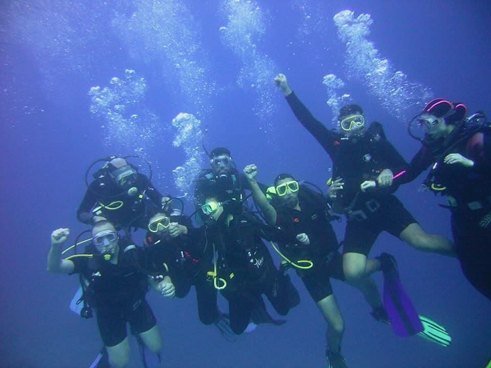 Group of seven divers underwater posing with fist pumps and peace signs, bubbles rising in deep blue water