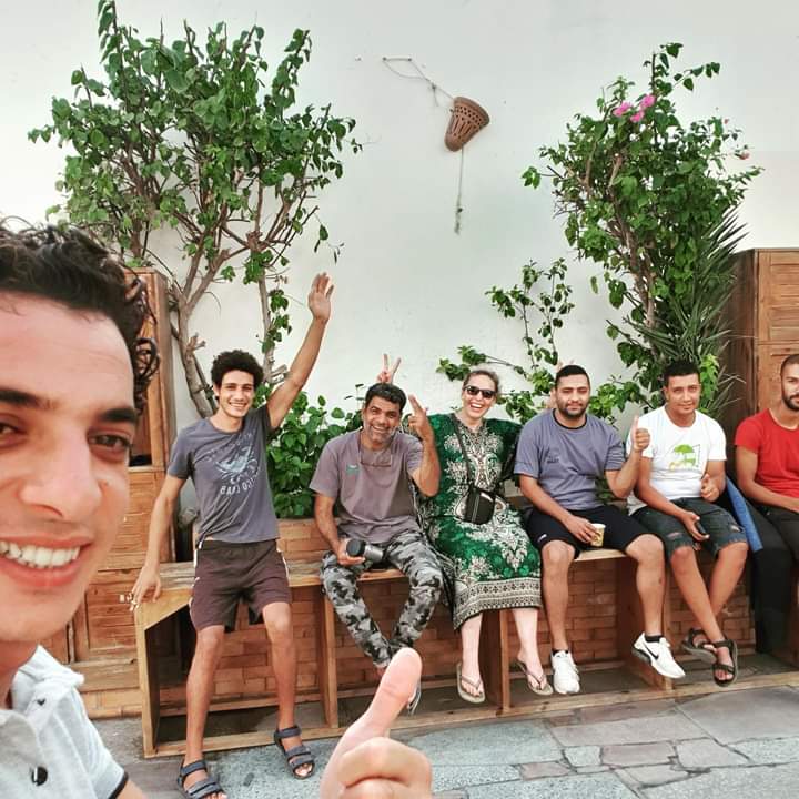 Group selfie of Osama with six friends and divers sitting on a wooden bench in a courtyard with green plants in Dahab