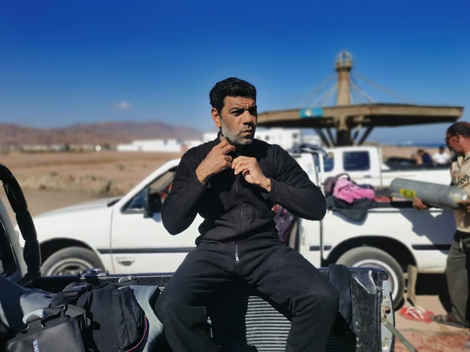 Osama sitting on the back of a pickup truck in his black wetsuit, zipping up, with desert landscape and dive tanks visible behind him