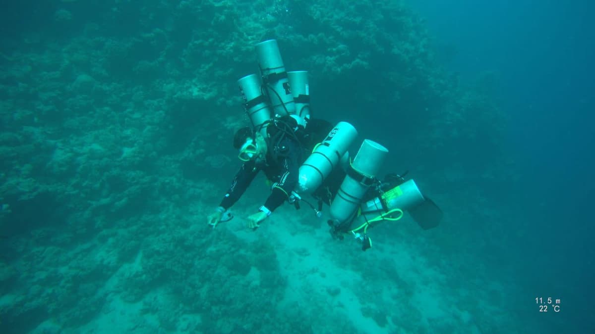 Osama diving underwater carrying seven tanks in a technical diving configuration, depth reading 11.5 meters at 22 degrees