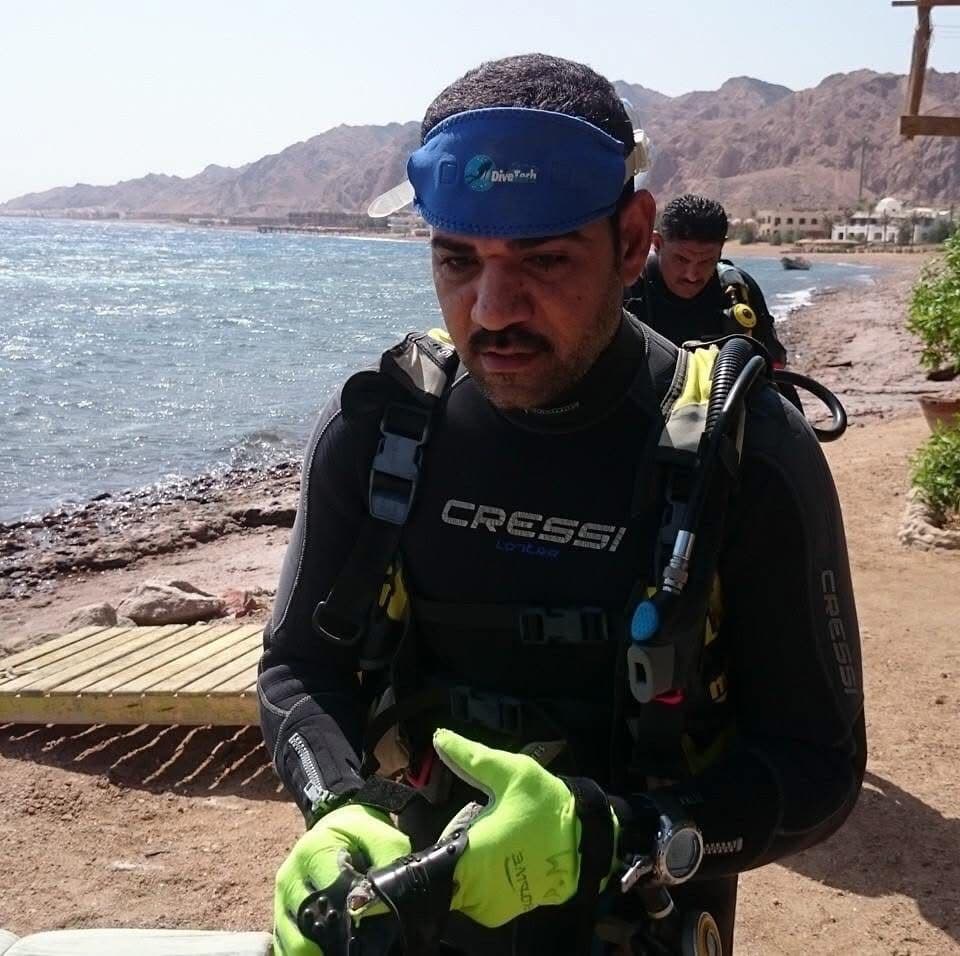Osama in full Cressi dive gear at Dahab shoreline preparing for a dive, Sinai mountains and Red Sea behind him