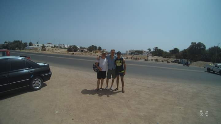 Three people standing on a wide desert road in Sinai, flat sandy landscape stretching to low buildings on the horizon, a dark car parked nearby, temperature reading 30 degrees