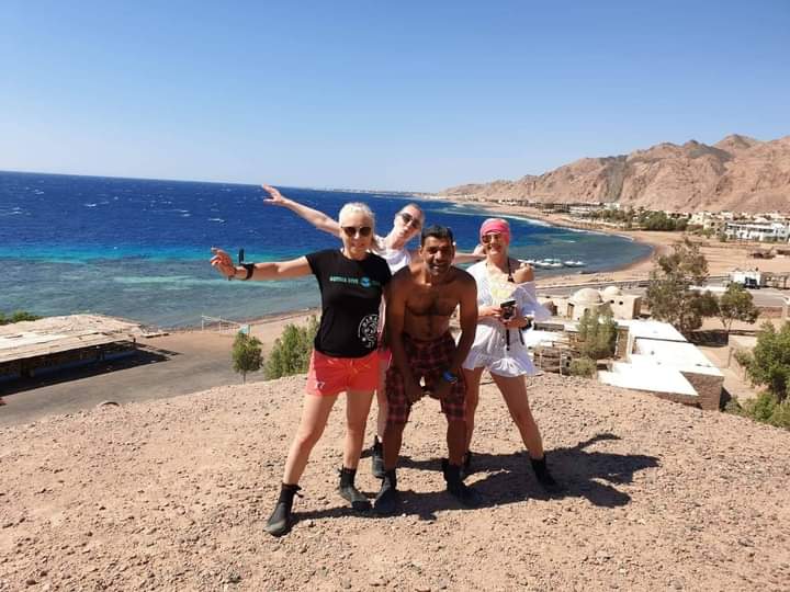 Four friends posing on a hilltop overlooking the Dahab coastline, turquoise reef visible below, red Sinai mountains stretching along the shore, one woman in a Bottle Dive Team shirt