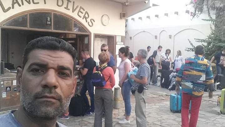 Osama taking a selfie at the entrance of Planet Divers dive center, a crowd of divers with bags and gear filling the doorway behind him, the curved Planet Divers sign overhead