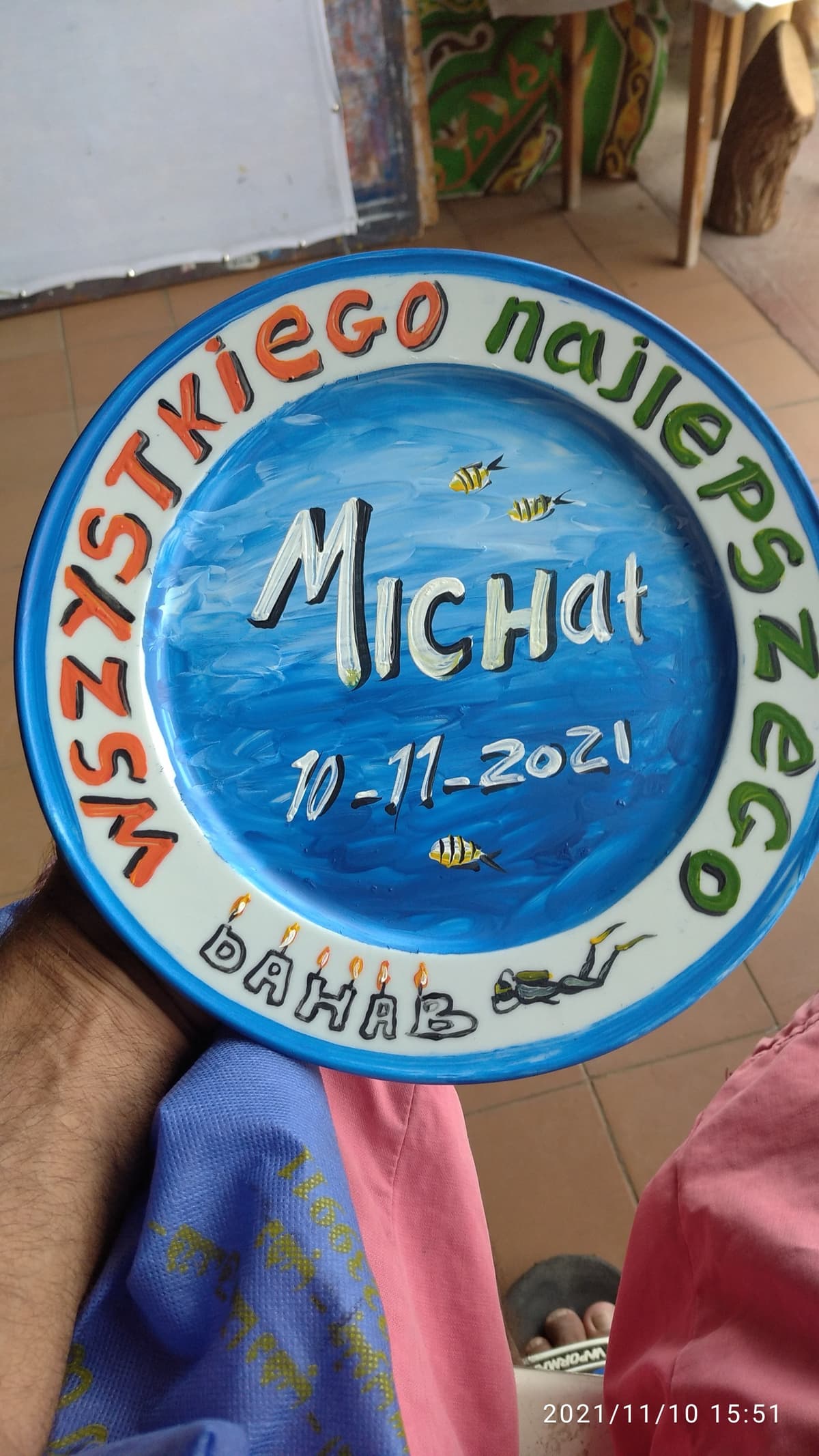 Hand-painted blue ceramic birthday plate reading Wszystkiego najlepszego Michal 10-11-2021 Dahab with painted fish, candles, and a diver, held over colorful Bedouin fabric