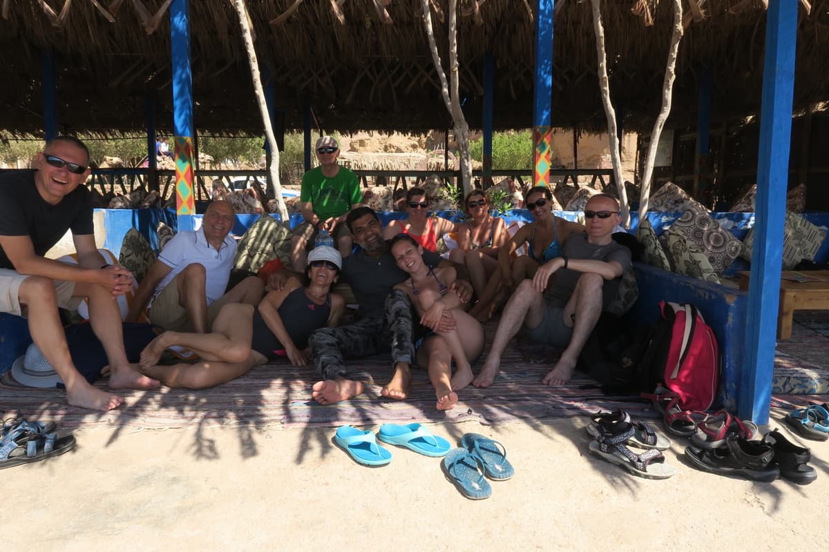 Group of about ten divers lounging barefoot in a colorful Bedouin beach shelter with blue wooden poles, woven mats and cushions, flip flops scattered in the sand, desert vegetation visible behind