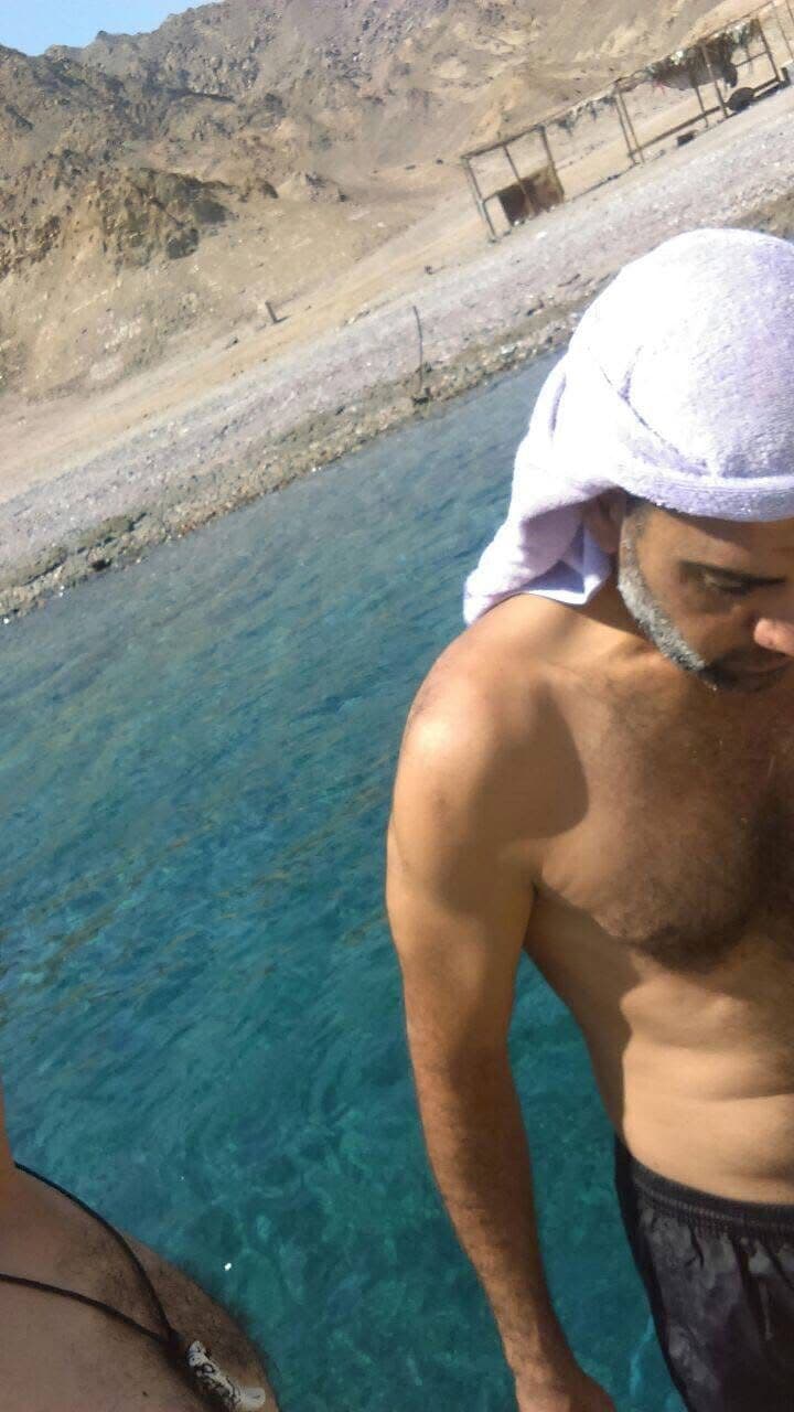 Close-up of Osama with a towel on his head looking down at the turquoise Blue Hole water from the cliff edge, barren Sinai mountain rising behind, Bedouin shelters visible on shore