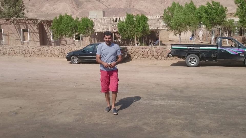 Osama standing alone in a dusty Sinai desert parking area wearing red shorts and a grey t-shirt, stone wall and trees behind, pickup truck and car parked nearby, mountains in distance