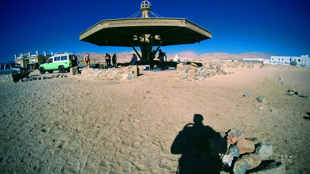 Wide-angle view of a large ornate metal dive site shelter in the middle of the Sinai desert, divers and vehicles gathered underneath, photographer's shadow stretching across the sand, 27 degrees