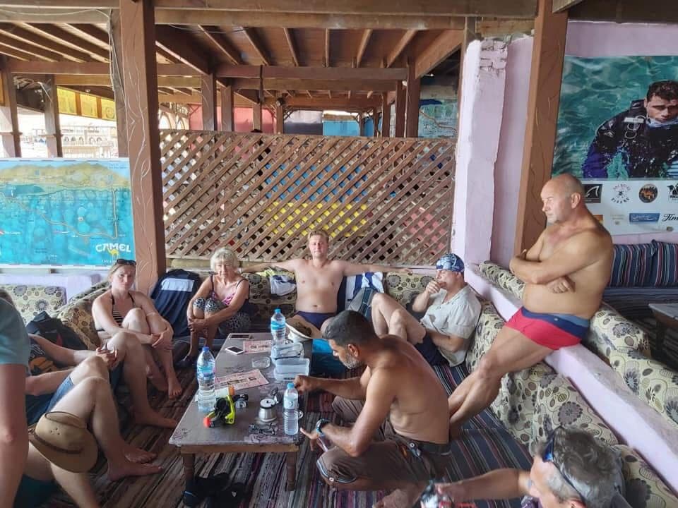 Group of seven divers relaxing in a Bedouin-style hut with cushions, a Camel Dive map and diver poster on the wall, lattice partition, water bottles and paperwork on a low table, Osama crouching at the table