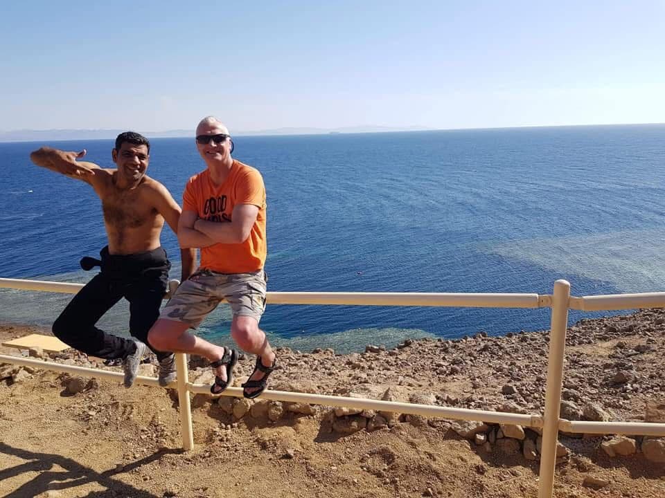 Osama shirtless in wetsuit bottoms and a friend in an orange shirt sitting on a metal railing above the Blue Hole, deep blue sea stretching to the horizon, rocky cliff beneath them