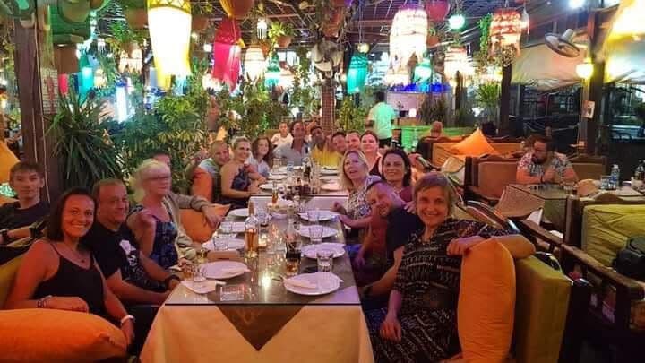 Large group of about twenty divers at a long dinner table at Shark Restaurant in Dahab, decorated with colorful hanging lanterns, tropical plants, and orange cushions, everyone smiling at the camera