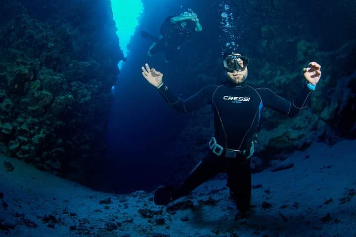 Osama kneeling on sandy bottom inside The Canyon wearing Cressi wetsuit, arms spread wide, another diver visible in the blue gap behind him