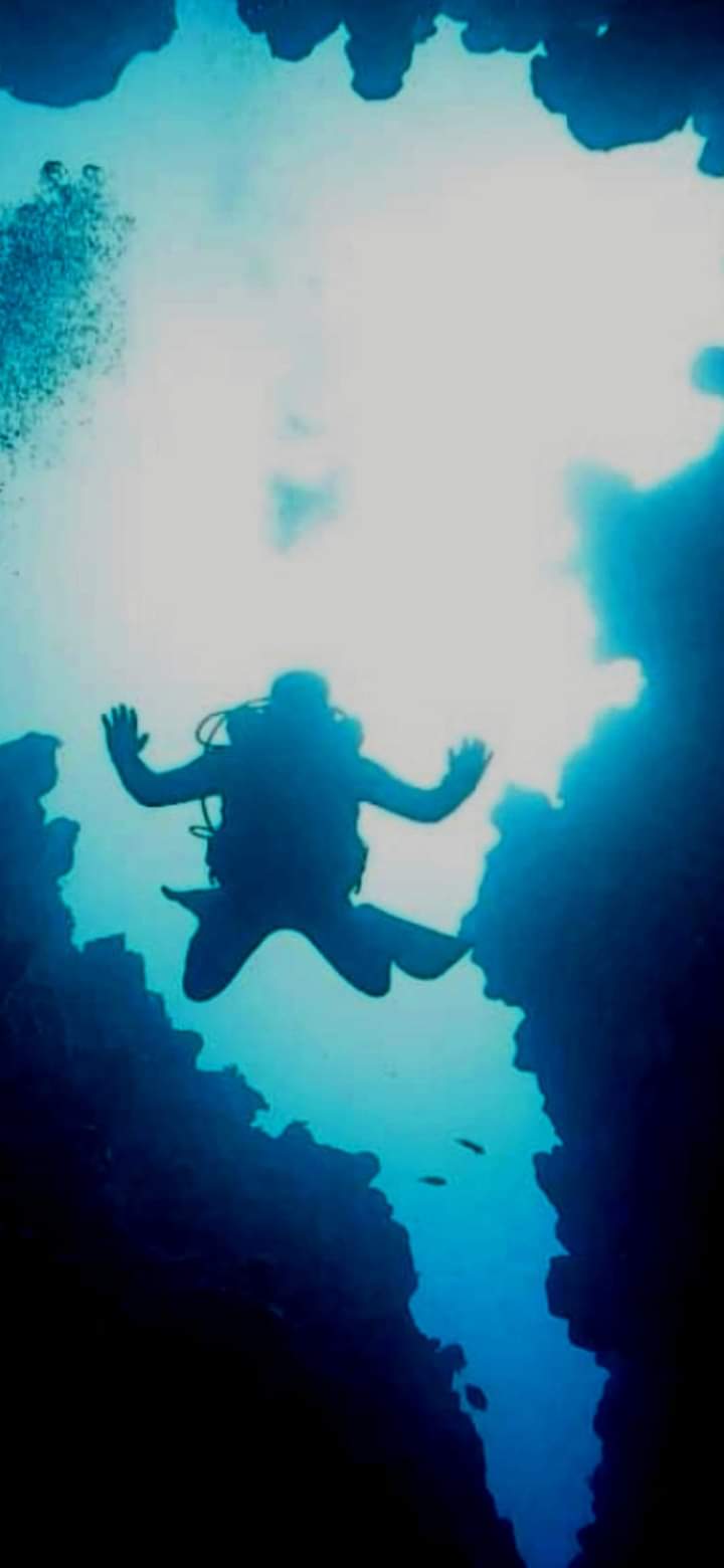 Diver silhouetted with arms spread wide inside The Canyon narrow gap, sunlight pouring through jagged rock walls from above, bubbles rising