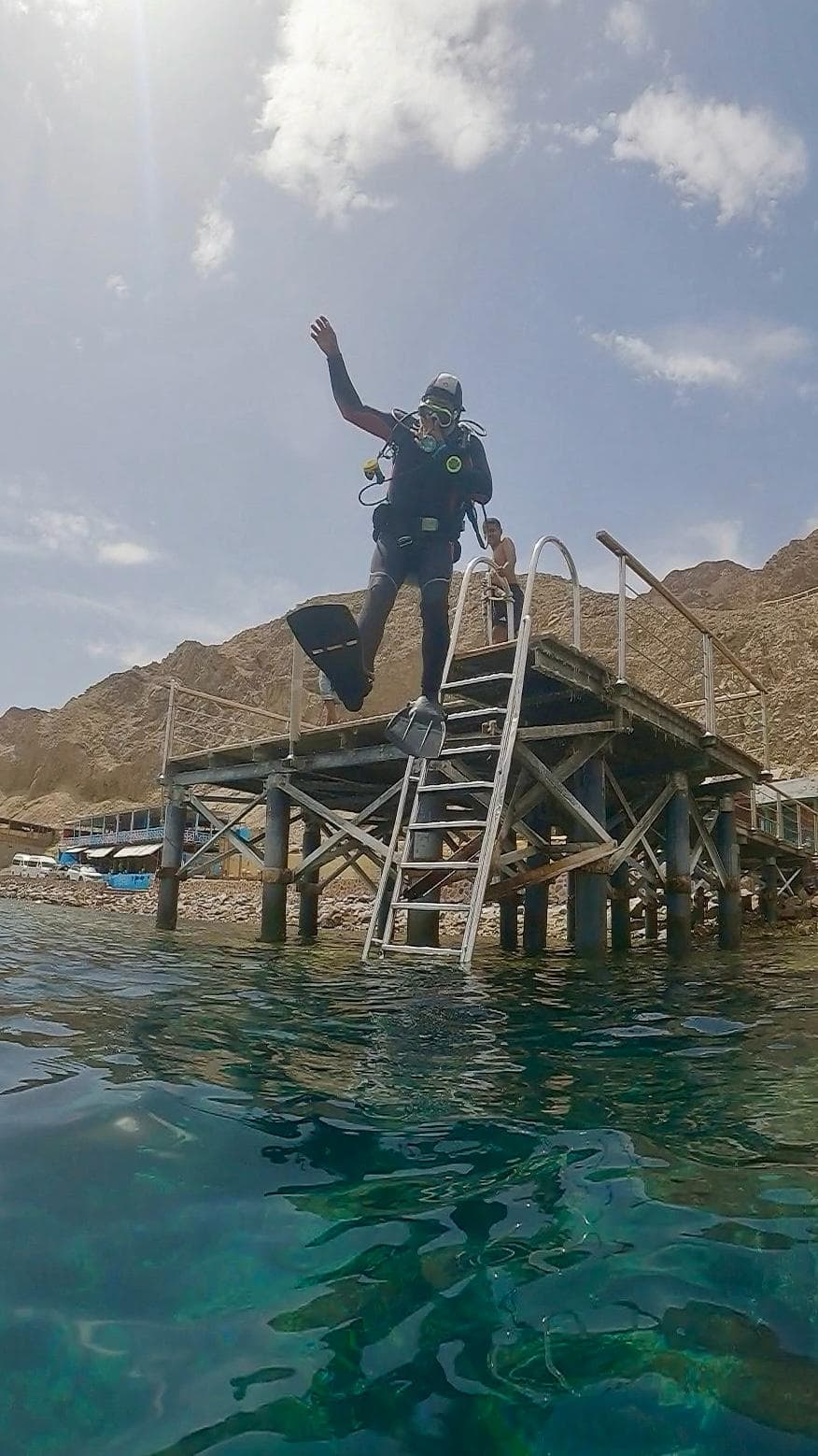 Diver mid-air doing giant stride entry off a wooden pier into turquoise water, desert mountains in background, ladder and shelters visible