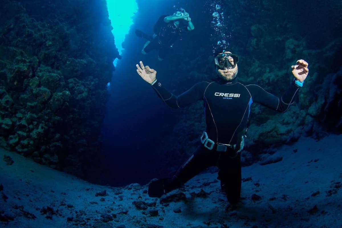 Osama in Cressi wetsuit kneeling on the sandy bottom of The Canyon, arms wide, blue light glowing through the narrow passage behind, another diver hovering above