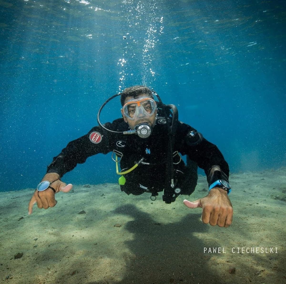 Osama hovering just above the sandy bottom in shallow crystal clear water doing double shaka hand signs, wearing dive gear, professional photo by Pawel Ciecheslki