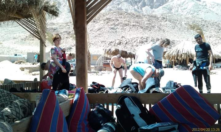 Divers preparing gear under a palm-thatched wooden shelter at a desert dive site, BCDs and regulators spread on Bedouin cushions, Sinai mountains behind, April 2012