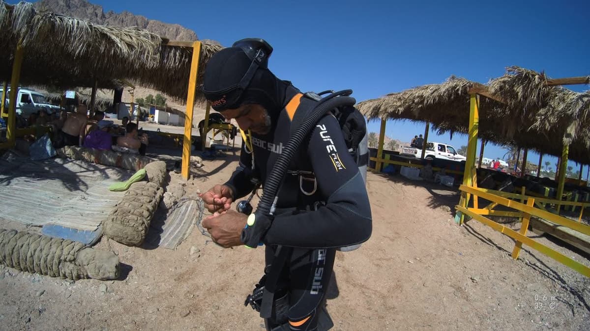 Osama in full ScubaSub BCD and Aqualung regulator checking his gear at a beach dive site, palm-thatched shelters and desert cliffs behind, bright blue sky