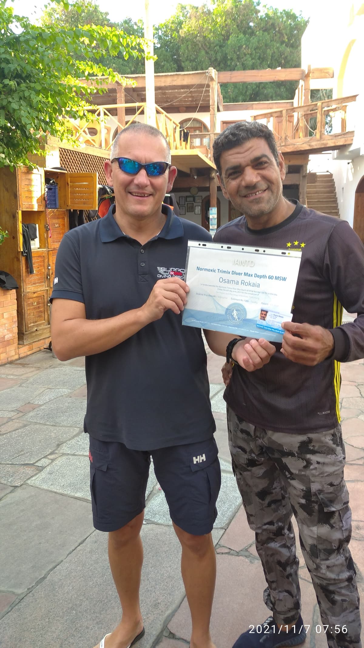 Osama smiling as his instructor Andrzej Kruczkowski in a Helly Hansen polo and blue sunglasses presents the IANTD Normoxic Trimix Diver certificate, wooden dive center courtyard in background