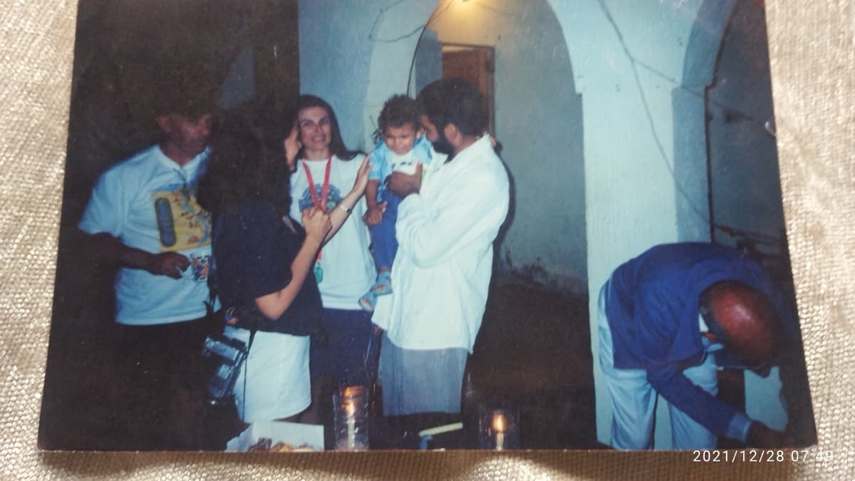 Faded vintage photograph of a young man in a white galabiya holding a small child while a woman smiles beside them, blue arched doorway and candlelight visible, another man in a printed t-shirt to the left