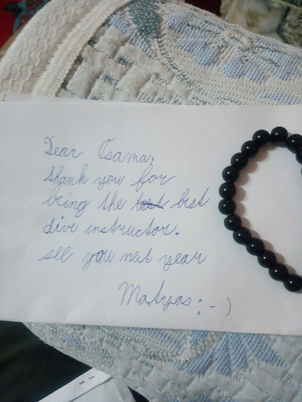 Handwritten note on white paper reading Dear Osama thank you for being the best dive instructor see you next year Matyas with a smiley face, a black bead bracelet placed beside it on lace fabric