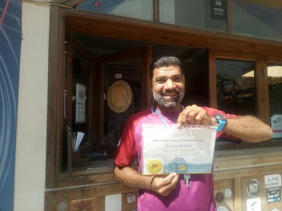 Osama in a red and pink shirt grinning while holding up his IANTD Advanced Rec Trimix Diver Max Depth 51 MSW certificate, PADI and other dive agency stickers on the window behind him