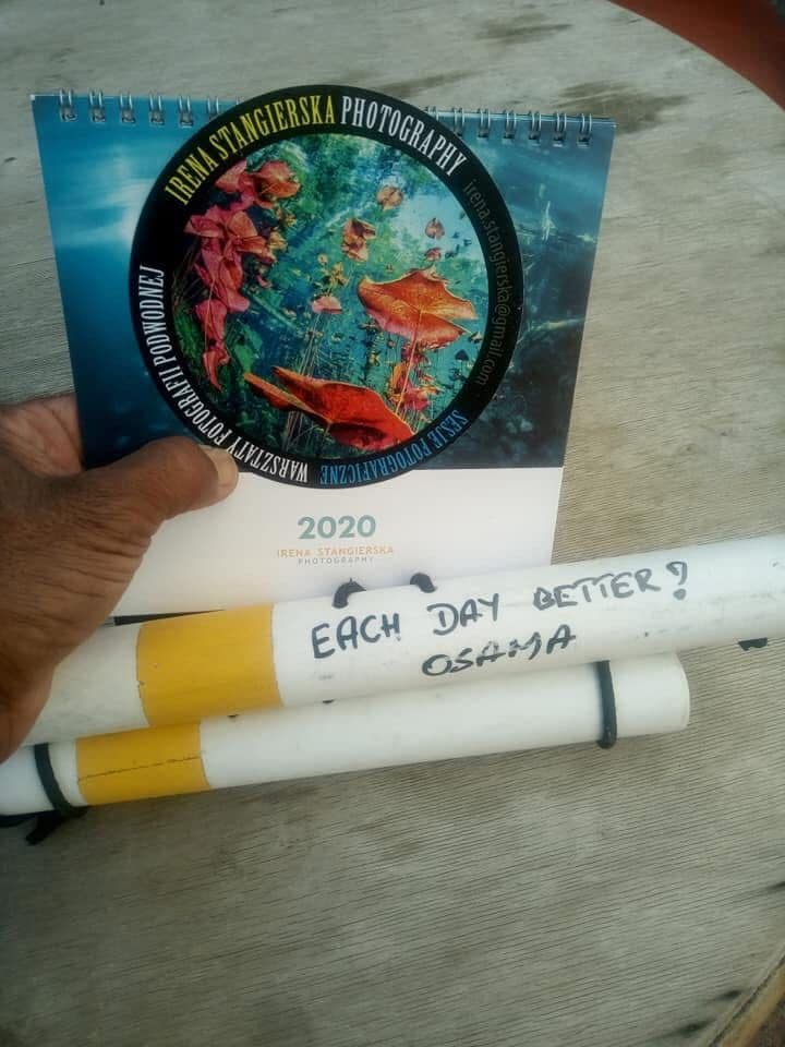Osama's hand holding an Irena Stangierska Photography 2020 underwater calendar with Polish text and a white dive marker reading Each Day Better Osama, on a wooden table