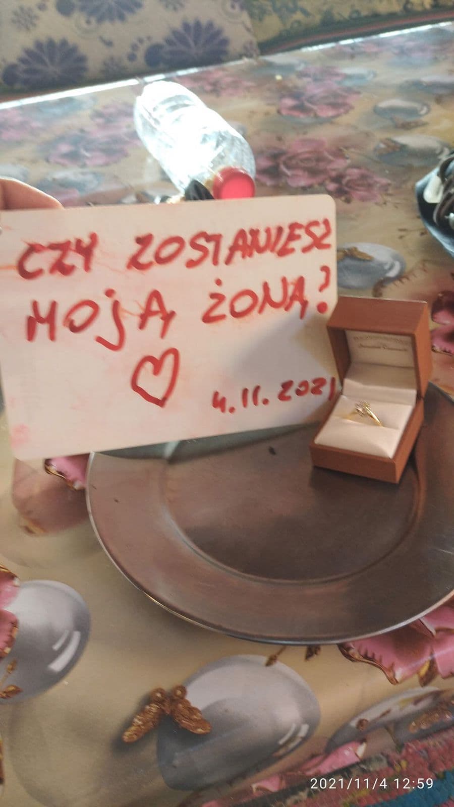 Waterproof proposal card with red handwritten Polish text Czy zostaniesz moja zona with a heart and the date 4.11.2021, a brown ring box with gold engagement ring beside it on a floral tablecloth