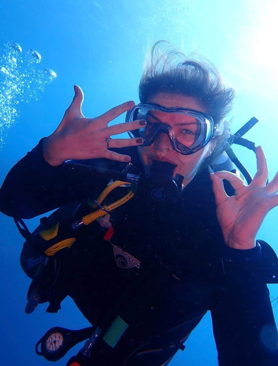 Kasia underwater showing her engagement ring on her left hand while making an OK sign with her right, hair floating in the blue water, bubbles rising, sunlight streaming from above, dive gauges visible