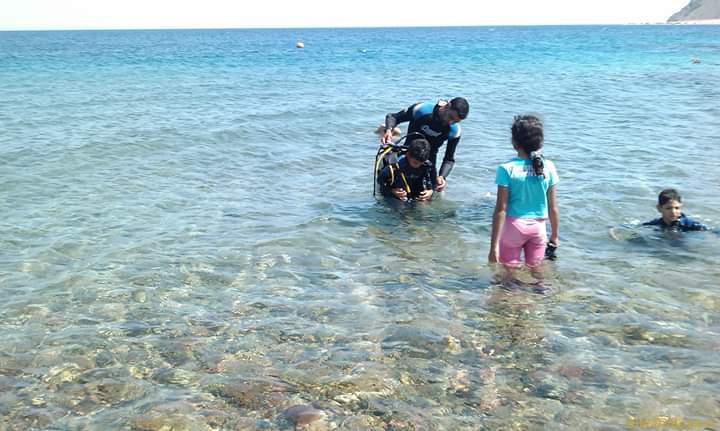 Osama in dive gear helping a student in crystal clear shallow water while a girl in turquoise shirt and a boy watch from nearby, Dahab coastline behind