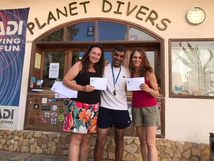 Osama standing between two smiling women holding their PADI certification envelopes in front of the Planet Divers entrance, PADI banner visible