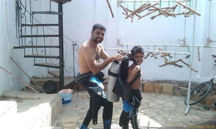 Osama smiling while helping a young boy put on a scuba tank at a courtyard in Dahab, spiral staircase and coral decorations on the wall, May 2012