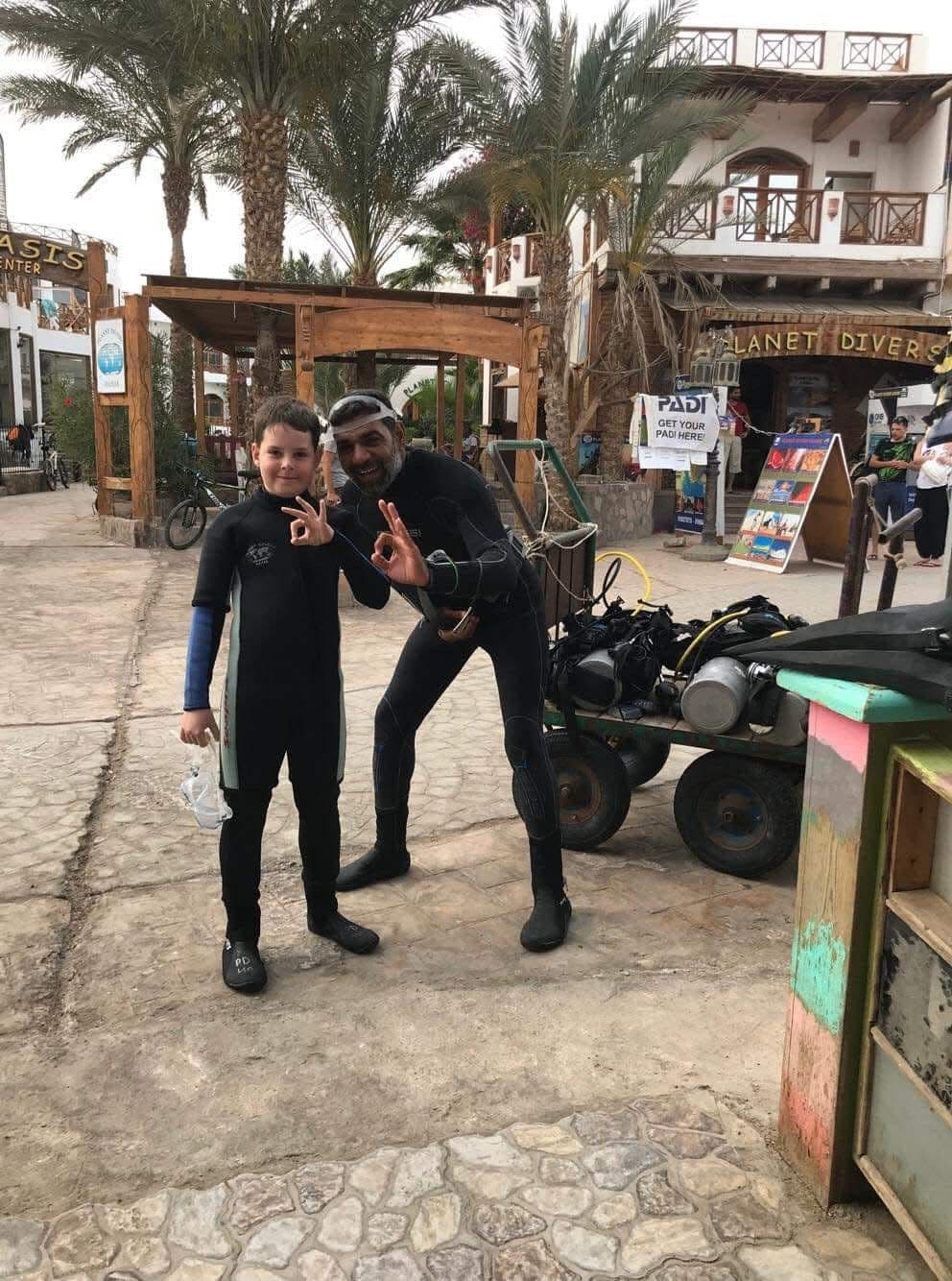Osama in black wetsuit crouching beside a smiling boy in a wetsuit, both doing OK signs, Planet Divers sign and PADI poster visible behind them, palm trees overhead