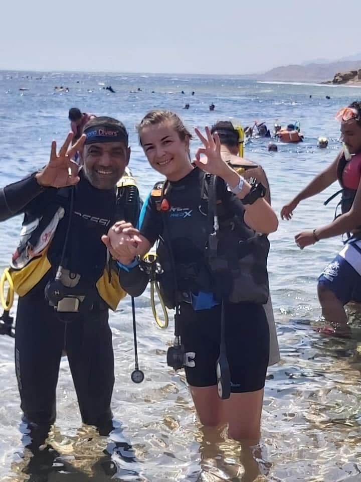 Osama wearing Cressi gear and Pro Divers mask strap standing in shallow water with a young female student, both smiling and doing OK signs, snorkelers and other divers in the busy background