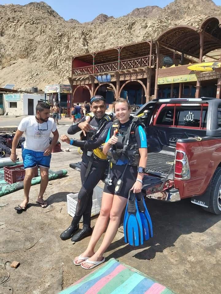 Osama in Cressi gear giving thumbs up standing with a young female diver holding blue fins at the Blue Hole parking area, desert cliffs and Bedouin shelters behind, red pickup truck nearby