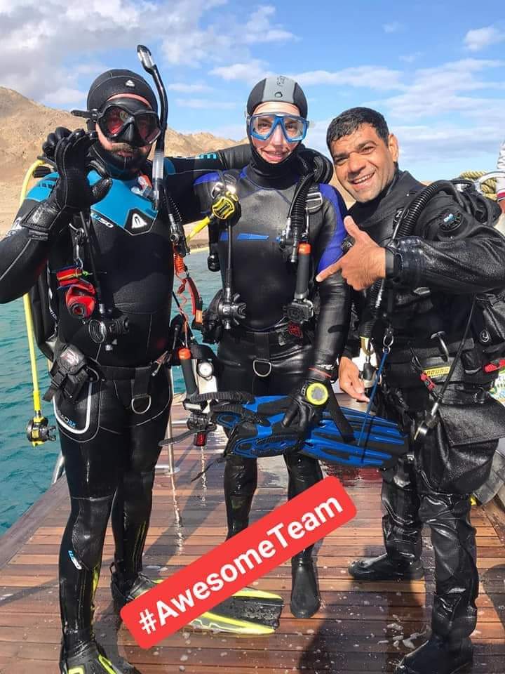 Three divers in full technical dive gear posing on a wooden boat dock with turquoise water and Sinai mountains behind, AwesomeTeam hashtag overlay