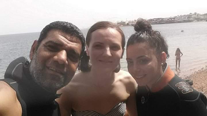 Osama in dive gear taking a beach selfie with two young women, one in a striped top and one in an orange wetsuit top, sandy beach and calm sea behind them