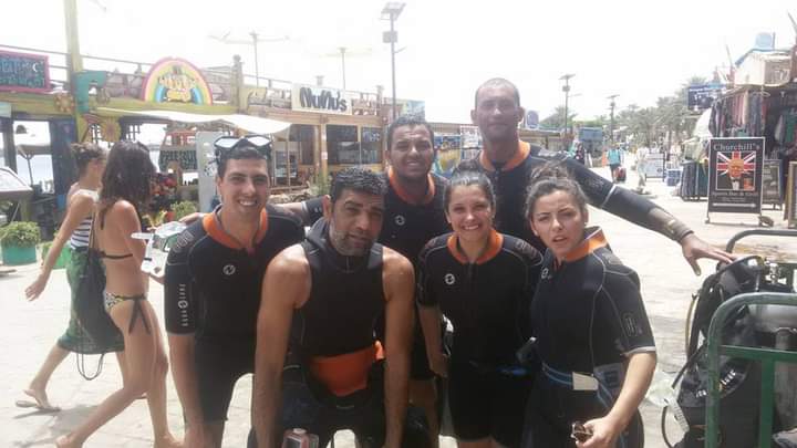 Five divers in matching black and orange wetsuits posing on the Dahab promenade, Churchill's bar and colorful shops behind them, Osama in the center