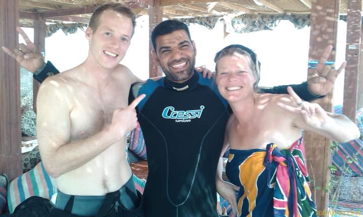 Osama in Cressi wetsuit with a young man giving thumbs up and a woman throwing rock horns under a Bedouin beach shelter, colorful cushions behind them