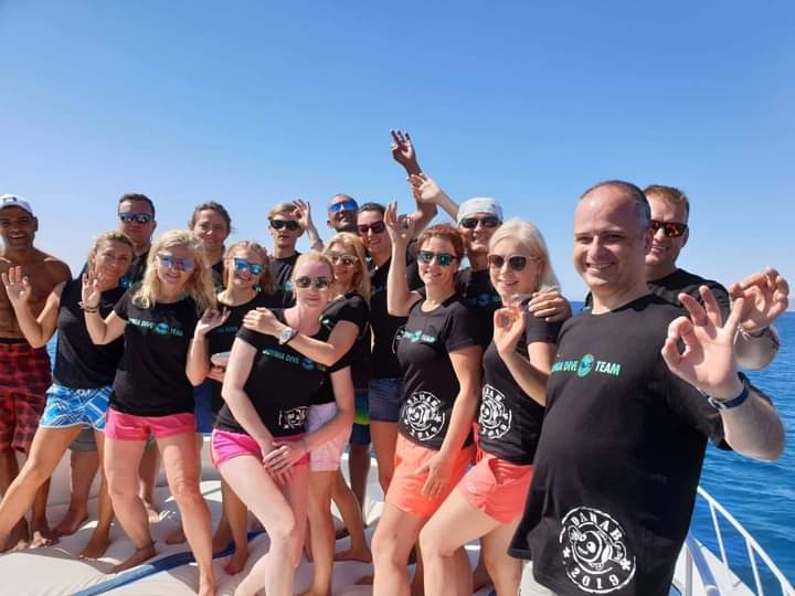 Fifteen divers in matching black Dahab Dive Team 2019 t-shirts posing on a dive boat, waving and giving OK signs, blue sky and open sea behind them