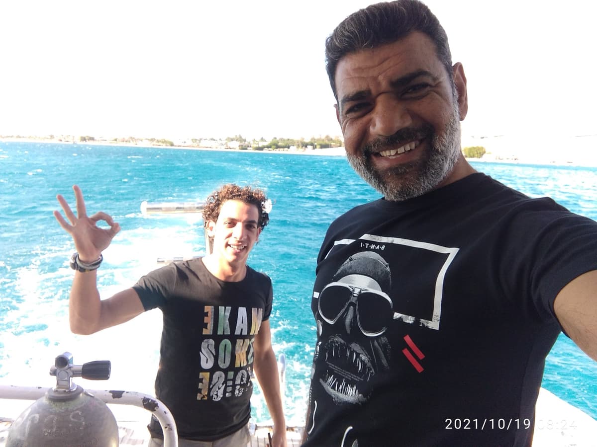 Osama in a black t-shirt with a diver skull print taking a selfie with a curly-haired young man doing OK sign on a dive boat, turquoise Dahab coast behind them, dive tank visible