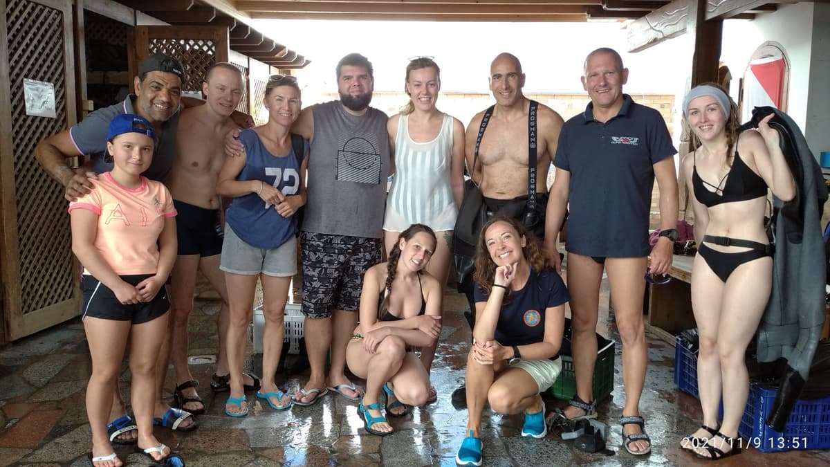 Osama with a group of ten divers and families of all ages posing under the dive center shelter, wet floor and dive flag visible, everyone smiling after a dive, November 2021