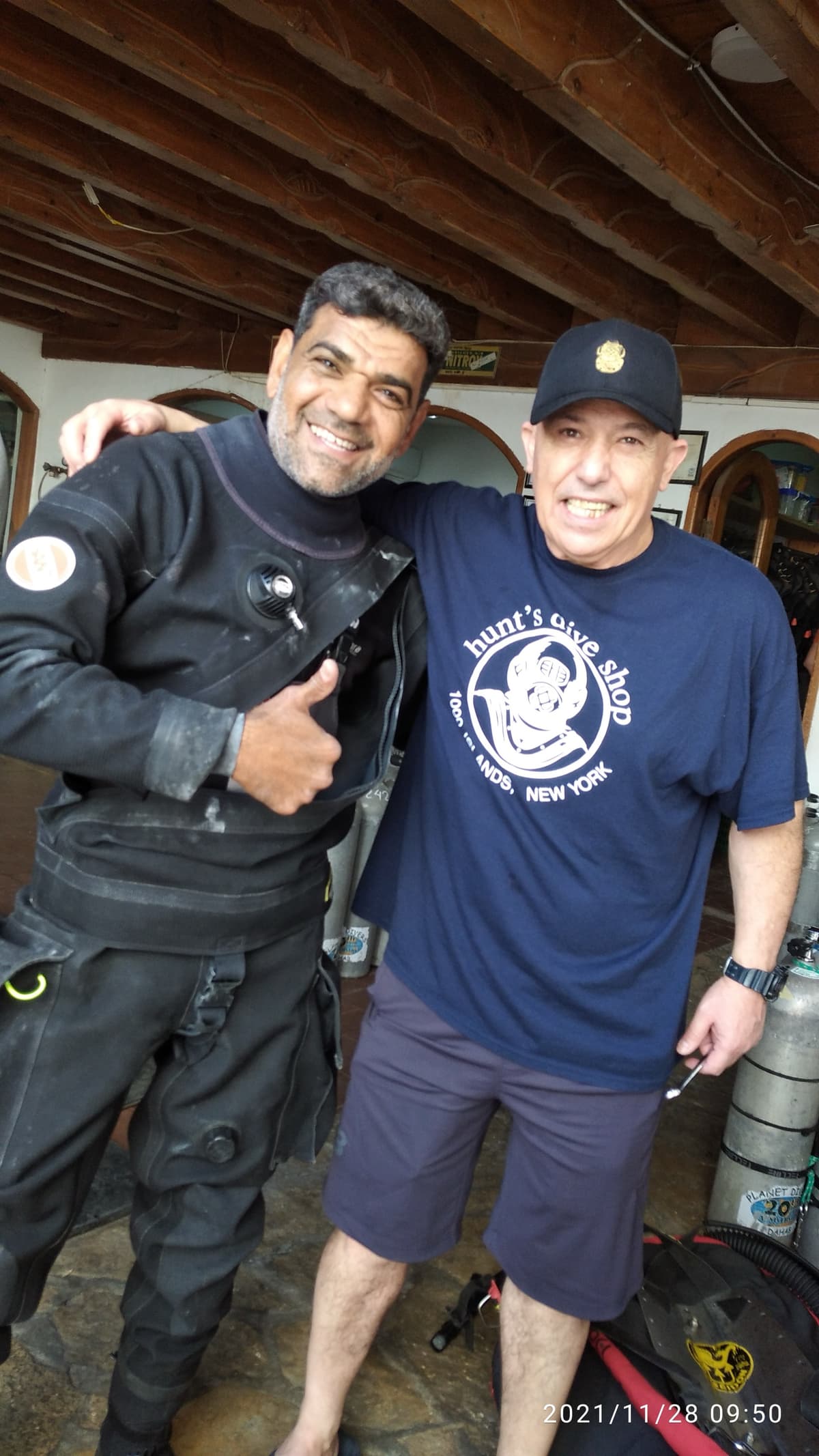 Osama in full black technical dive gear giving thumbs up next to Nuno Gomes wearing a Hunt's Dive Shop Andes New York t-shirt and black cap, tanks and wooden beams of the dive center behind them