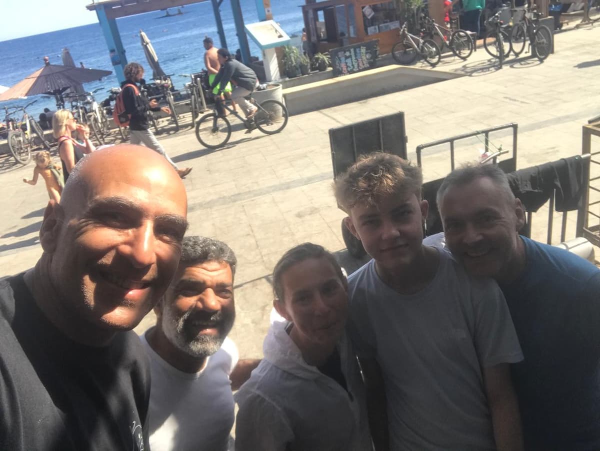 Osama taking a group selfie with four friends on the Dahab promenade, beach umbrellas and bicycles and the sea visible behind, bright sunny day
