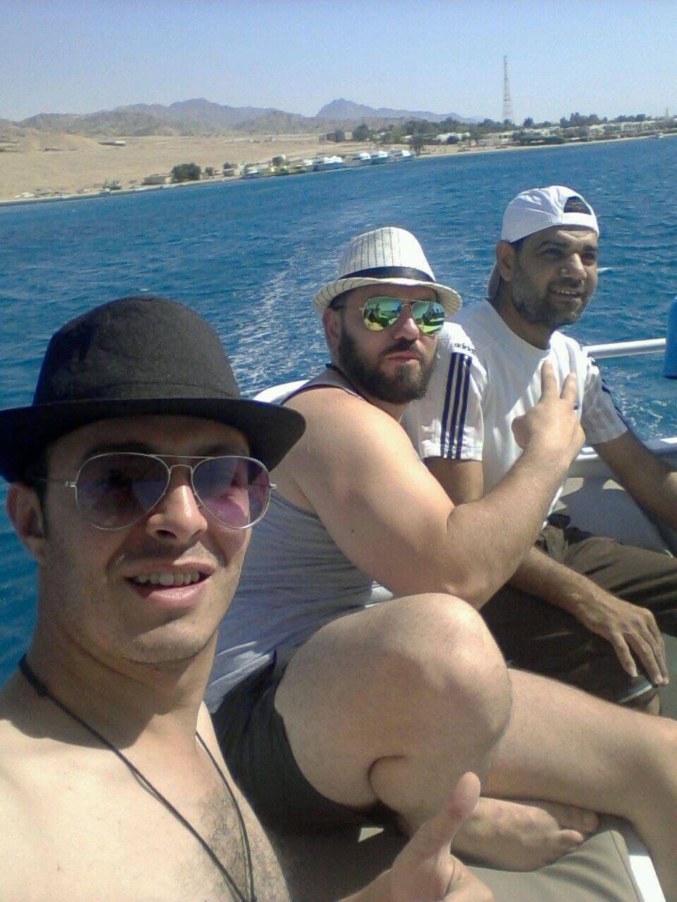 Three men selfie on a boat - one in aviators and a black hat, one in mirrored sunglasses and a striped bucket hat, one in a white cap - Sinai desert coastline and cell tower visible across the blue water