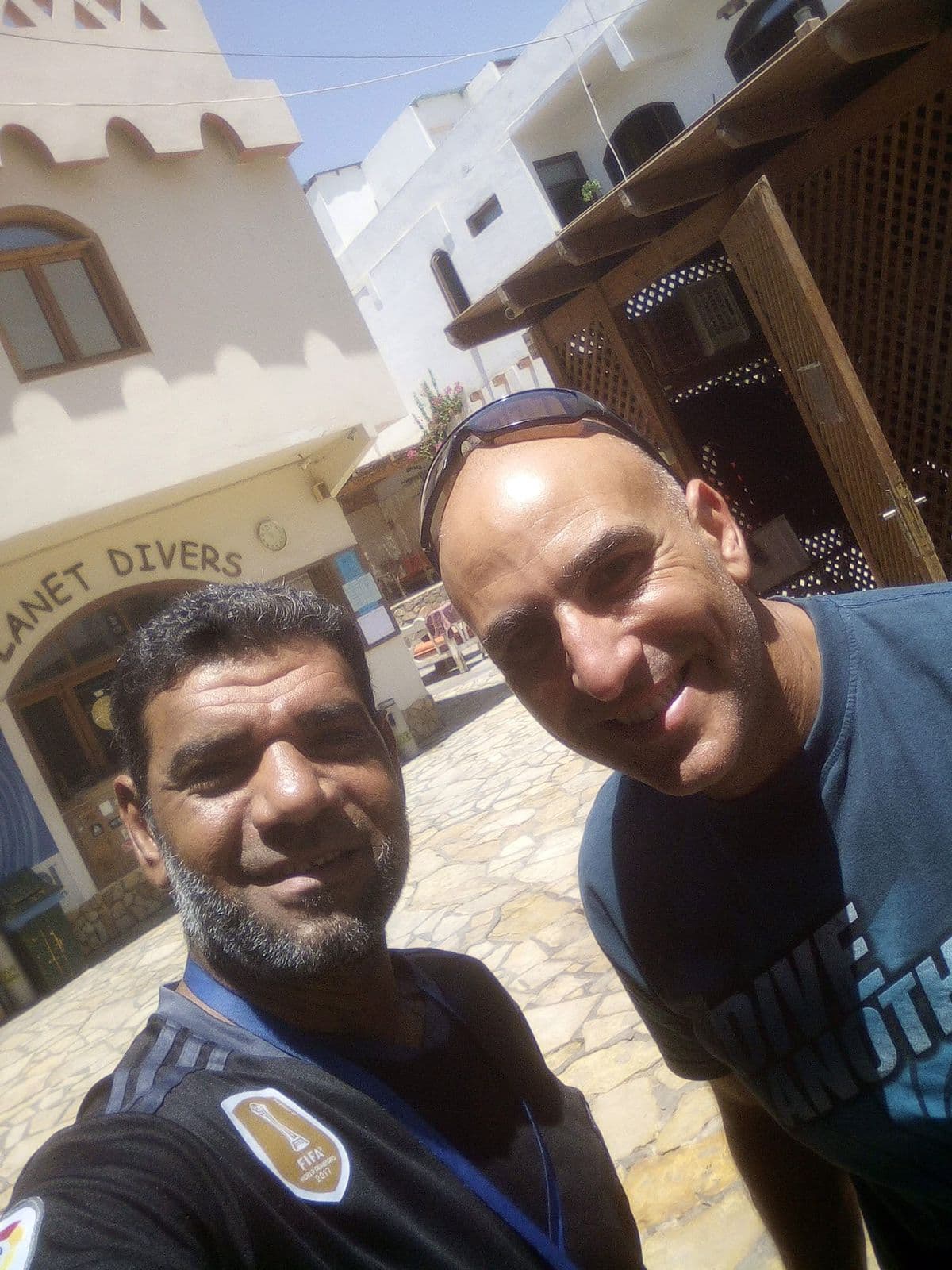 Osama taking a selfie with a friend wearing a Dive Instructor shirt in front of Planet Divers entrance, white Dahab buildings and wooden lattice balcony behind them