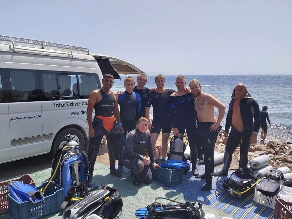 Eight divers in wetsuits posing by a white Dive Club International van at a rocky shore, Cressi and Salvimar gear visible, tanks and fins spread on a green mat, sea and cliffs in background