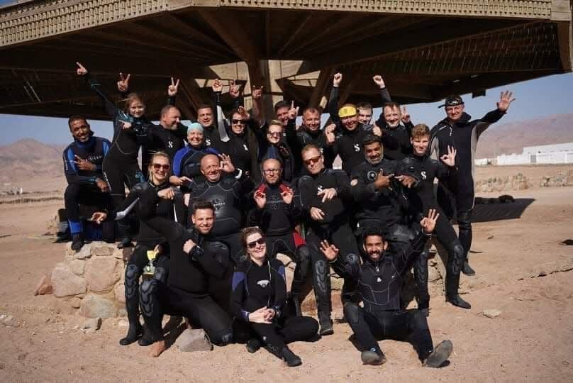 Over twenty-five divers in black wetsuits posing in rows under a large metal desert shelter, Scubapro logos visible, everyone throwing hand signs, Sinai mountains and white buildings in the background